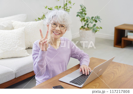 ノートパソコンの前でピースサインするシニア女性　リビングルーム 117217808