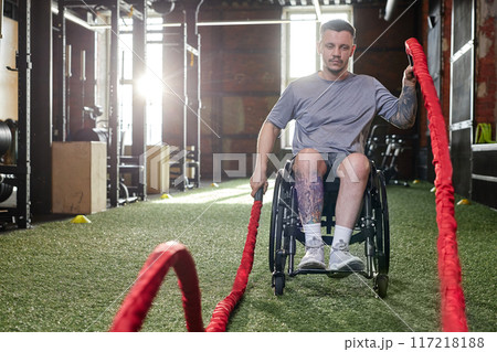 Front view full length portrait of young man with disability using wheelchair during strength training in gym with battle rope, copy space 117218188