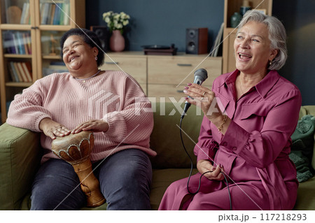 Candid portrait of two cheerful senior women playing music together and singing to microphone at home 117218293