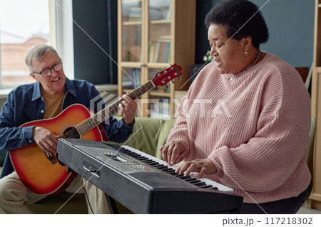 Side view portrait of Black senior woman playing piano and singing with senior man with guitar in background copy space Side view portrait of Black senior woman playing piano and singing with senior man with guitar in background copy space 117218302