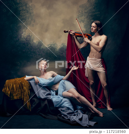 Man playing violin for young tender woman draped in blue fabric against vintage background. Sense of romance, creativity and admiration. Man playing violin for young tender woman draped in blue fabric against vintage background. Sense of romance, creativity and admiration. 117218560