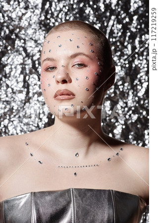 Vertical portrait of young woman wearing glittering makeup with gems looking at camera with confidence on metallic background Vertical portrait of young woman wearing glittering makeup with gems looking at camera with confidence on metallic background 117219259