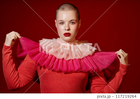 Fashion portrait of bald young woman posing in studio wearing red dress with ruffles and looking at camera 117219264