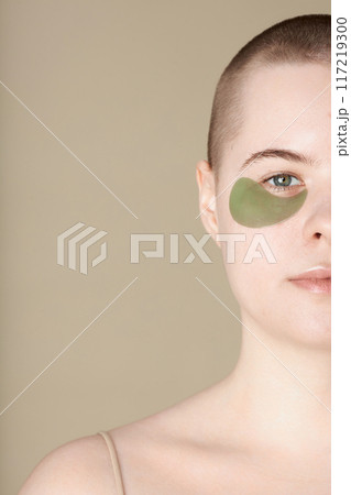 Minimal half portrait of youthful girl wearing moisturizing eye patch looking at camera on beige background copy space 117219300