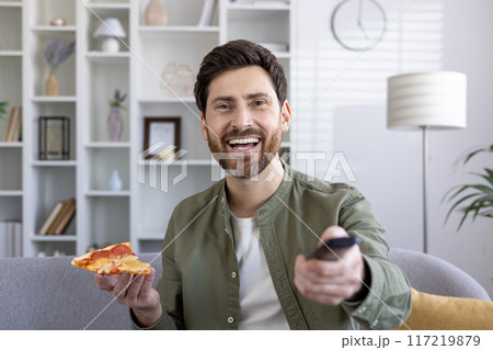 Cheerful man holding slice of pizza while watching TV at home. Smiling adult relaxing on sofa with remote control and delicious snack, enjoying leisure time 117219879