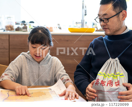 ハザードマップを見て自分の家の場所を確認している娘とそれを見守る非常持ち出し袋を抱えたお父さん 117220099