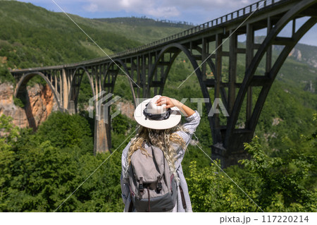A girl in a hat stands against the backdrop of a beautiful arched bridge over the Tara River, a popular tourist attraction in Montenegro. A girl in a hat stands against the backdrop of a beautiful arched bridge over the Tara River, a popular tourist attraction in Montenegro. 117220214