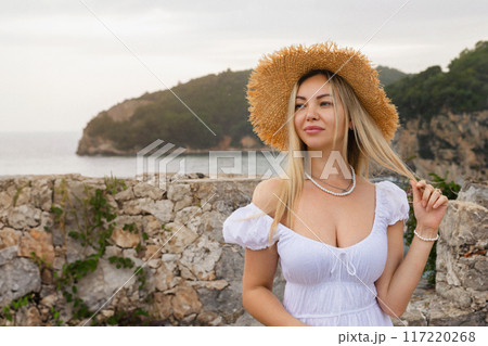 Charming fantasy woman in a hat posing for the camera. Young spectacular woman in a summer dress with bare shoulders in the background on a sunny summer beach 117220268