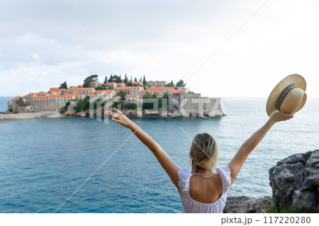 girl tourist in a hat looks from a top on the island of Sveti Stefan in Montenegro and the sea girl tourist in a hat looks from a top on the island of Sveti Stefan in Montenegro and the sea 117220280