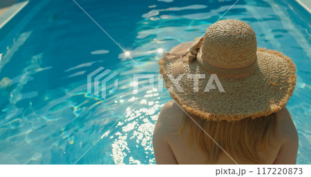Young woman in a hat sitting by the pool, admiring the sun reflections on the water 117220873