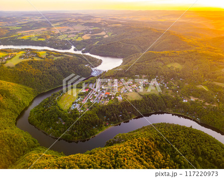 The sun sets over the tranquil Slapy Water Reservoir, casting golden hues across the landscape. Lush green hills frame the winding Vltava River, while a quaint village nestles along the water. 117220973