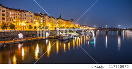 A view of the Rasin Embankment and Palacky Bridge in Prague, Czechia at dusk. The bridge is lit up, and the city lights reflect in the water. There are boats on the river, and the sky is a deep blue. 117220976