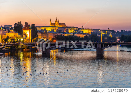 A view of Prague Castle illuminated at dusk as seen from a bridge over the Vltava River. The city lights reflect on the calm waters, creating a serene and picturesque scene. Prague, Czechia 117220979