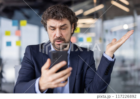 Confused businessman in suit looking at cell phone in modern office setting. Man appears perplexed or frustrated while holding smartphone, likely dealing with unexpected news or information. 117226892