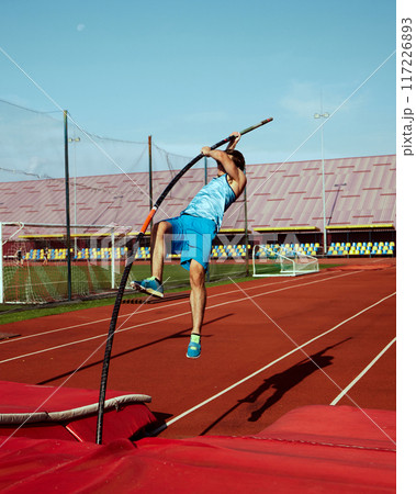 High jump practice in action, with focused and strong pole vaulter, competitive man in blue uniform training at outdoor stadium 117226893