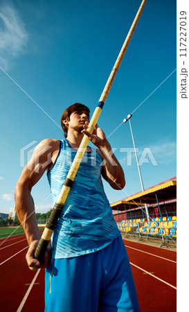 Pole jump athlete, focused young man in blue uniform standing with pole, showing reading for training session at open air stadium Pole jump athlete, focused young man in blue uniform standing with pole, showing reading for training session at open air stadium 117227019