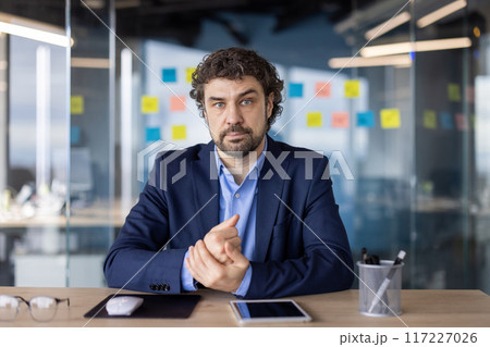 Confident businessman in blue suit sitting at desk in modern office with digital tablet and office supplies. Professional corporate setting with sticky notes on glass wall in background. 117227026