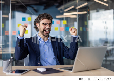 Businessman in suit celebrating success at office desk with laptop, showing excitement and joy. Professional environment with modern decor, highlighting achievement, success, and satisfaction. Businessman in suit celebrating success at office desk with laptop, showing excitement and joy. Professional environment with modern decor, highlighting achievement, success, and satisfaction. 117227067