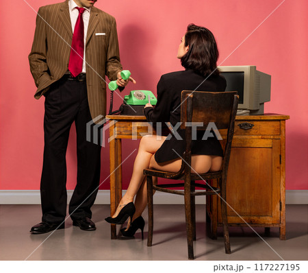Suited man, boss holding rotary phone and points while woman in heels and blazer sits at a desk with retro computer against oink background. Suited man, boss holding rotary phone and points while woman in heels and blazer sits at a desk with retro computer against oink background. 117227195