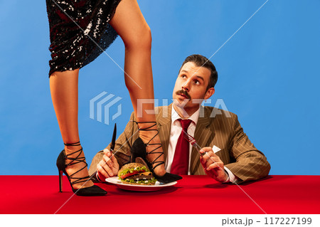 Stylish woman in heels pours liquid into glass from above, while man in suit sits at red table with burger, looking up in surprise. 117227199