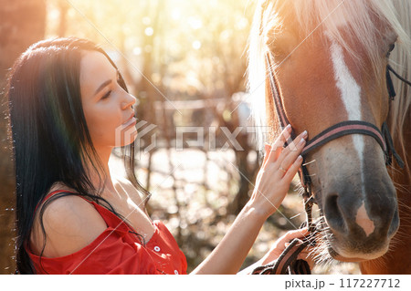 Portrait of a Young beautiful brunette woman in a red dress stroking a horse on the face. Sunlight Portrait of a Young beautiful brunette woman in a red dress stroking a horse on the face. Sunlight 117227712