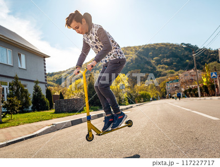 A teenage boy performs a trick on a scooter, Bouncing up on it. In the background, an empty street and a view of the mountain. Side view. Concept of extreme sports, tricks and youth activity A teenage boy performs a trick on a scooter, Bouncing up on it. In the background, an empty street and a view of the mountain. Side view. Concept of extreme sports, tricks and youth activity 117227717