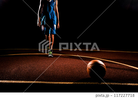 Basketball. A teenage boy in a blue sports uniform walks confidently towards a basketball ball lying on the Playground. Black background. Concept of sports games 117227718