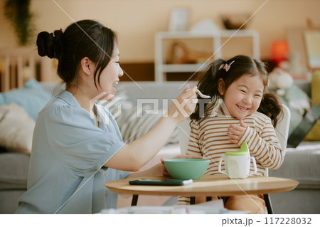 Asian mom smiling widely while feeding her little daughter in nursery Asian mom smiling widely while feeding her little daughter in nursery 117228032