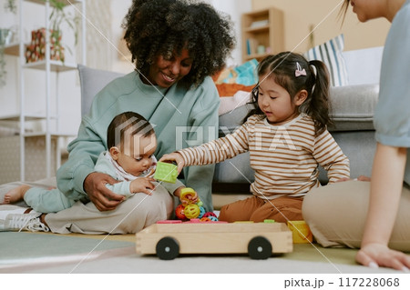 Little Asian girl giving green brick to baby boy who sitting in African American mothers arms, they sitting on comfortable carpet surrounded by toys Little Asian girl giving green brick to baby boy who sitting in African American mothers arms, they sitting on comfortable carpet surrounded by toys 117228068