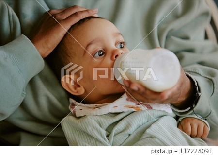Medium close up of male infant wearing bib and eating formula from baby bottle while caring hand of his mother patting his head Medium close up of male infant wearing bib and eating formula from baby bottle while caring hand of his mother patting his head 117228091