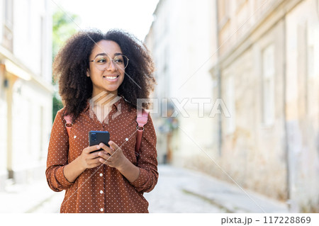 Woman tourist with curly hair and glasses strolling through a sunny city street, enjoying her vacation. She is holding a smartphone and wearing a brown shirt with a pink backpack. 117228869