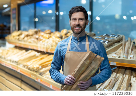 Bakery employee smiling while holding fresh baguettes. Bread section in background with various types of loaves. Concept of friendly customer service and fresh bakery products Bakery employee smiling while holding fresh baguettes. Bread section in background with various types of loaves. Concept of friendly customer service and fresh bakery products 117229036