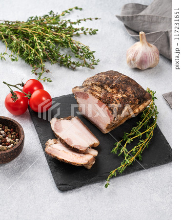 Baked pork belly sliced on a black slate board on a light background with fresh tomatoes and herbs. 117231651