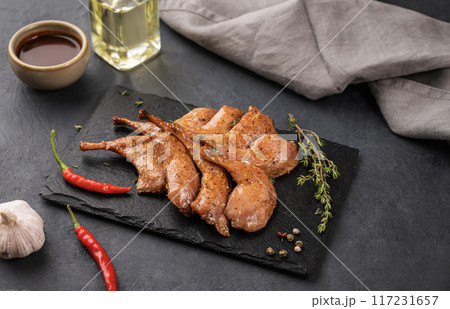 Grilled BBQ quail legs in marinade and spices on black slate with fresh herbs, oil Grilled BBQ quail legs in marinade and spices on black slate with fresh herbs, oil 117231657