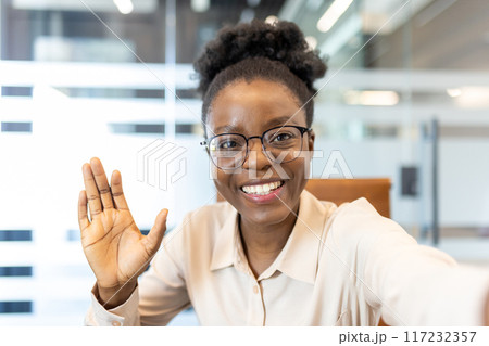 Smiling woman with glasses waving at camera during video call in office. Happy professional greeting while looking at the camera using phone. Concept of virtual meetings and remote communication. Smiling woman with glasses waving at camera during video call in office. Happy professional greeting while looking at the camera using phone. Concept of virtual meetings and remote communication. 117232357
