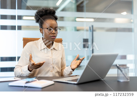 Young african american businesswoman using laptop for video call in office. Serious expression with hand gestures communicates important discussion or presentation over phone. Young african american businesswoman using laptop for video call in office. Serious expression with hand gestures communicates important discussion or presentation over phone. 117232359