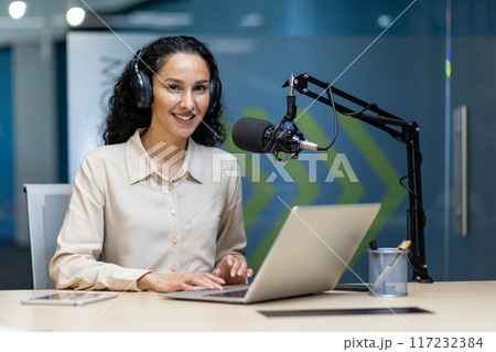 Smiling woman wearing headphones recording podcast using laptop and professional microphone in modern office. Engaged in content creation, broadcasting, online communication, media production 117232384