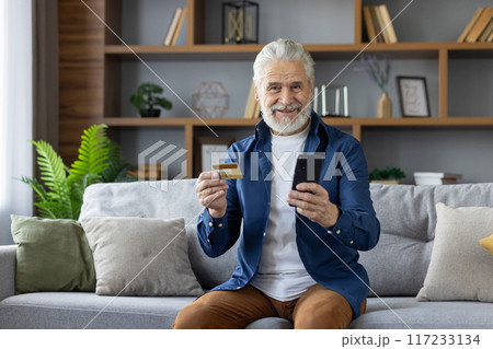 Portrait of a cheerful elderly Caucasian man holding a credit card and using a smartphone while sitting ,comfortably on a couch in a modern living room. 117233134