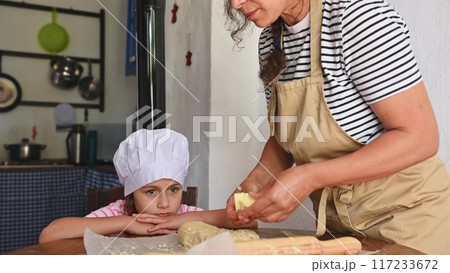 A mother and daughter spend quality time baking croissants together in a cozy kitchen. The little girl thoughtfully arranges baked goods while learning the art of cooking. A mother and daughter spend quality time baking croissants together in a cozy kitchen. The little girl thoughtfully arranges baked goods while learning the art of cooking. 117233672