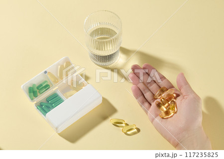 Creative idea for medication advertising pic, daily routine of medicine drinking with daily pill box, water glass and fish oil supplement held in hand palm. Space for promotional, high angle view 117235825