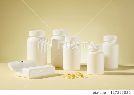 Several white mockup bottle without label neatly arranged on the table and featured on light yellow background, with a white pill box and some soft yellow supplement capsule flat lay on the table 117235826