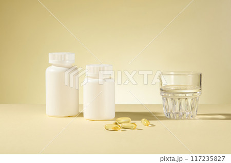 The photography at light yellow background features white unbranded supplement jars, a glass of water and yellow vitamin capsules. Front angle shot, blank space for adding text 117235827