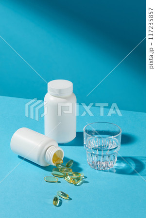 Yellow soft gel capsules spilling out of white bottle flat lay on blue table, next to an unbranded medicine bottle and a glass of water. Vitamin and supplement concept photo, high angle shot Yellow soft gel capsules spilling out of white bottle flat lay on blue table, next to an unbranded medicine bottle and a glass of water. Vitamin and supplement concept photo, high angle shot 117235857