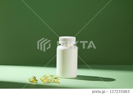Frontal photo taken on a green background with a streak of sun shining through an unlabeled white medicine bottle displayed in center, next to some flat lay supplement capsules. Advertising space 117235863
