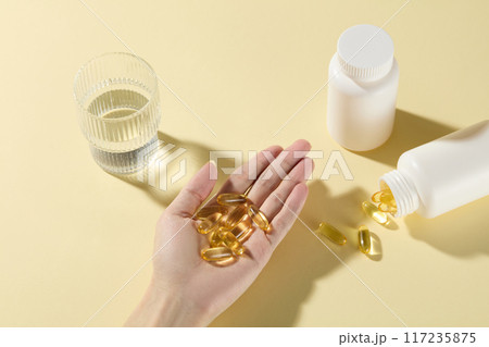 Medicine drinking preparation scene with a female hand holding yellow supplement capsules in palm, glass of water and unbranded white medicine bottles placed around. Top view, copy space for text 117235875