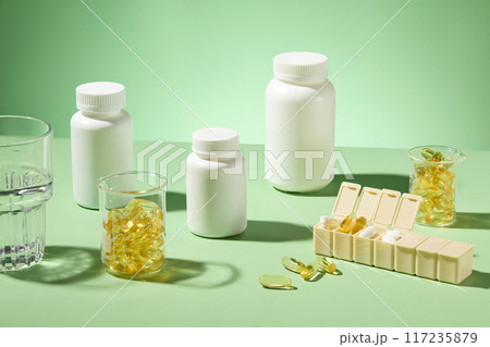 Multiple unbranded white medicine jars placed on green table with yellow supplement capsules contained in glass beaker and daily pill container, glass of water place on the edge. High angle view 117235879