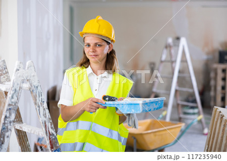 Woman house painter in yellow vest and hardhat in room being renovated 117235940