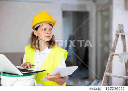 Female foreman in a protective helmet and a yellow vest checks the execution of repair work using laptop 117236032