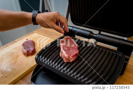 Chef at the kitchen preparing beef steaks on the home electric grill Chef at the kitchen preparing beef steaks on the home electric grill 117238543