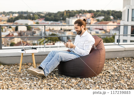 Man relaxing on rooftop with smartphone and laptop on bean bag Man relaxing on rooftop with smartphone and laptop on bean bag 117239883